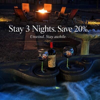A hot tub with water flowing, a bottle of wine and two glasses. Behind it is a fire pit. The words say Stay 3 nights. Save 20%. Unwind, Stay awhile.