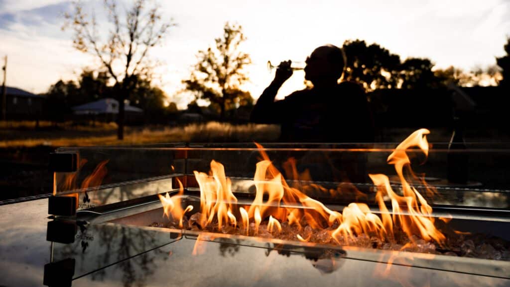 Silhouette of a person drinking from a glass beside a glowing fire pit at sunset outside.
