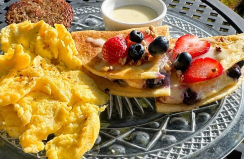 A glass plate on an outside table with scrambled eggs, blueberry and cheese quesadillas, a white dipping sauce, and sausage