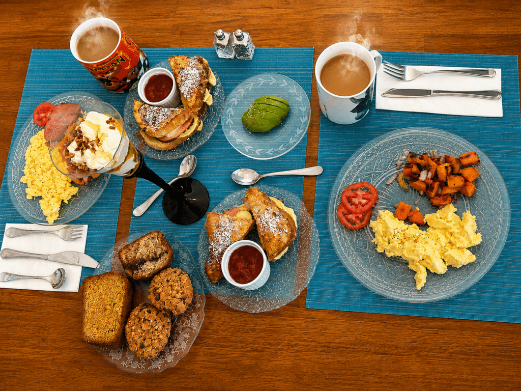 A breakfast spread of scrambled eggs, skillet sweet potatoes, avocado, steaming coffee, baked goods, and jam.
