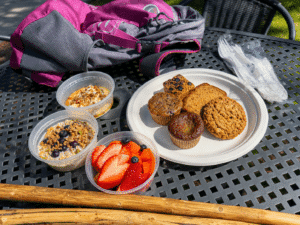 Various containers with fruit, overnight oats and yogurt beside a plate of baked goods, a backpack, and walking sticks.