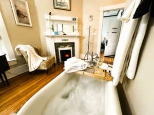 A bathroom with tub, fireplace, robe, towels. The tub is filled with bubbles and has a wine glass.