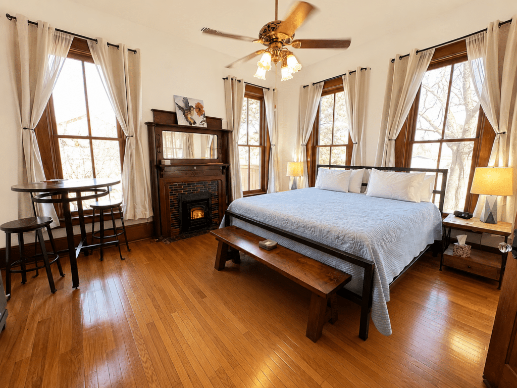 A large, sunlit room with 3 windows, a late 1800 fireplace and mantle, shining wood floors, a large bed. a bench, table and chairs.