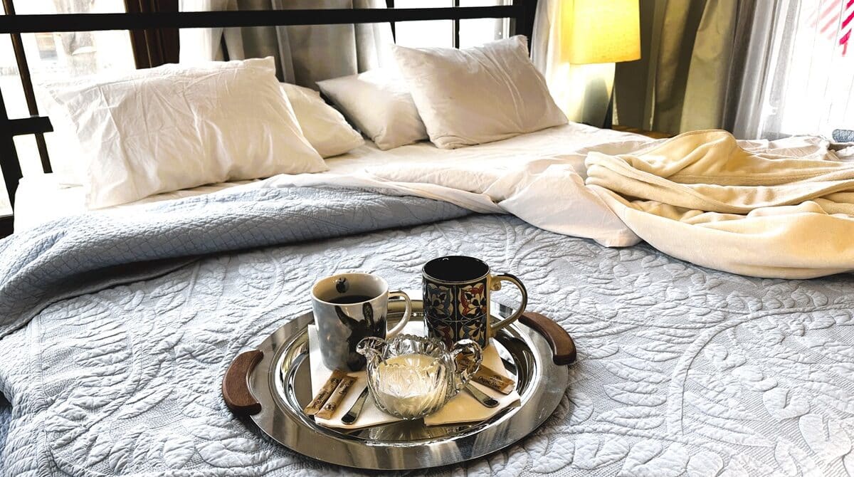 a coffe tray a bed with windows behind the bed. The tray has coffee, cream sugar and spoons