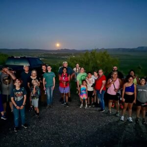 A group of people at twighlight with a full moon rising behind them.