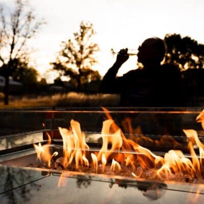 Picture of an outdoor firepit in low light or at sunset with a person behind it in silouette drinking from a champagne flute.