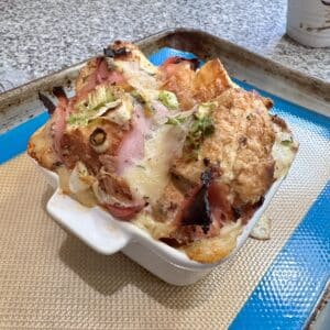 A croissant-based bread pudding with ham, onions, and cheese, puffing up out of the baking dish.