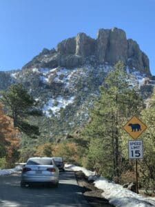 A photo taken from a car on a road with two cars in front and a snowy mountain in the distance.