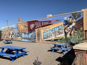 A colorful mural across the side of a building that depicts different aspects of Alpine - A Sul Ross banner, a postcard that says Greetings from Alpine Texas, Dan Blocker, Mariachis, and more.