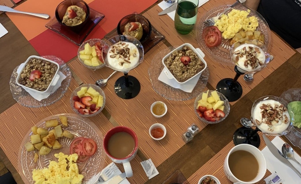 A table set with various dishes - yogurt, eggs, baked goods, fresh fruit, coffee, jams, baked apples, and more