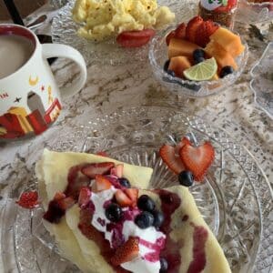 a plate of crepes with fruit on top, coffe, a bowl of fruit, and a plate of scrambled eggs.