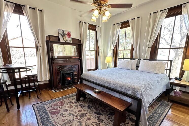 Sunlit room with king bed, 4 windows, large fireplace and mantle, bend, ceiling fan and light, nightstands, cafe table and stools