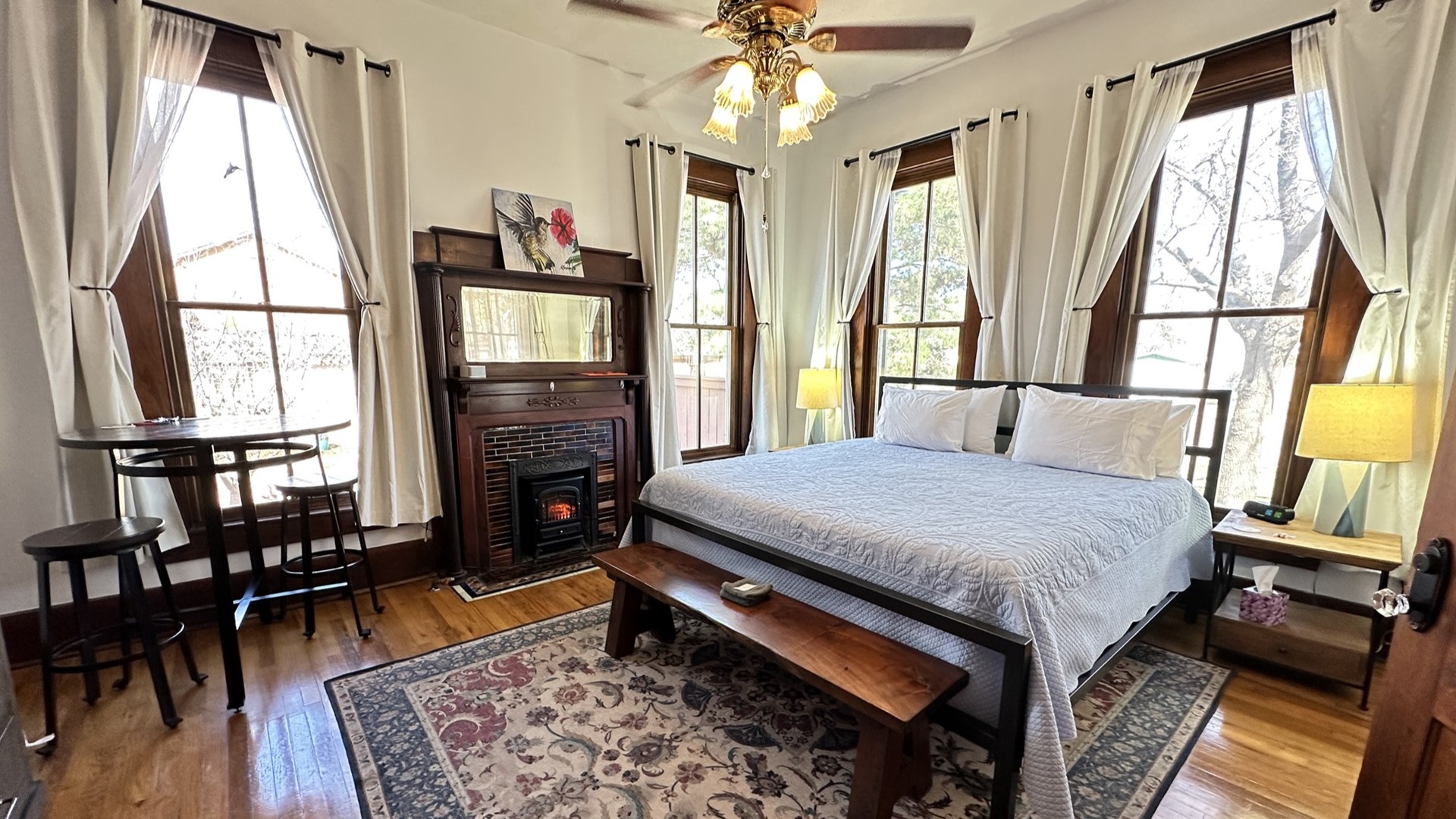 Sunlit room with king bed, 4 windows, large fireplace and mantle, bend, ceiling fan and light, nightstands, cafe table and stools