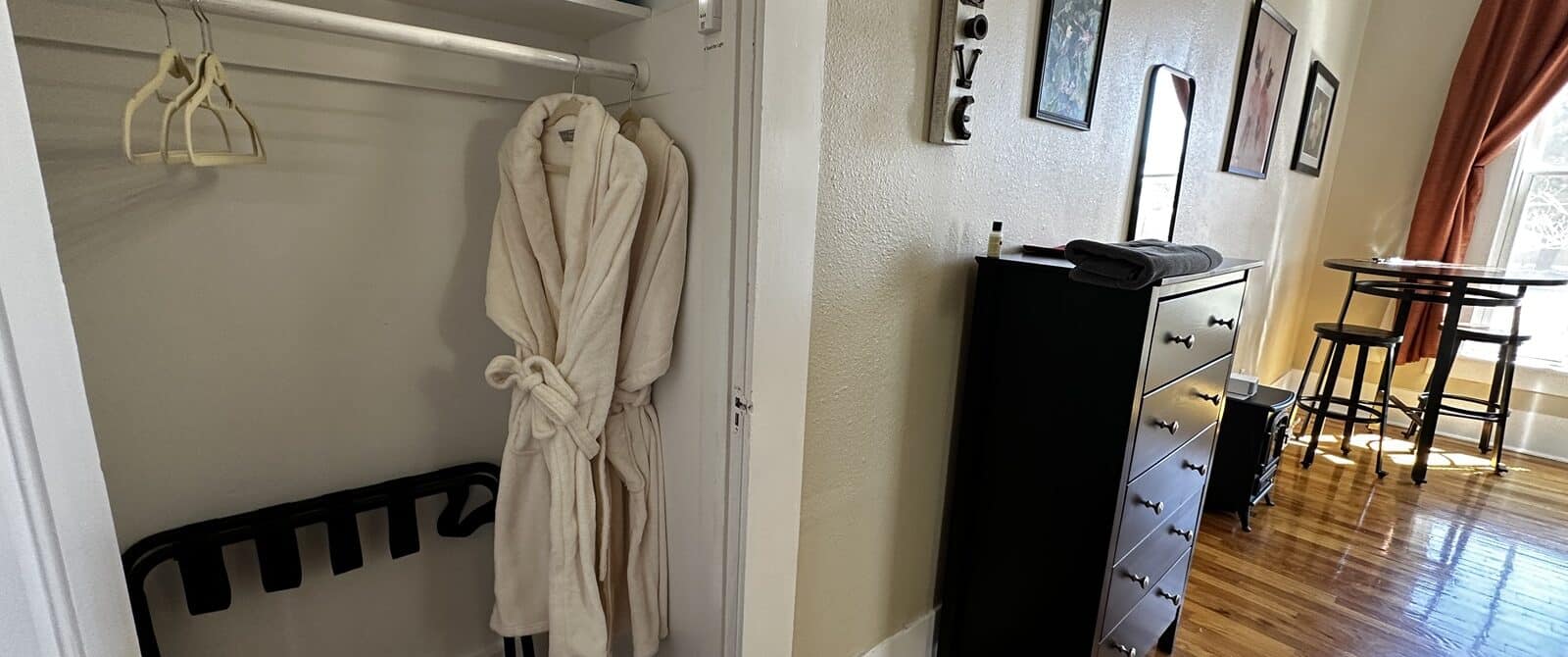 interior view of a bedroom showing a closet with two robes and a blanket, a dresser, and art.