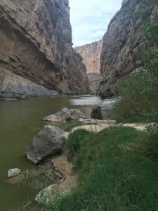 Towering canyon walls flank a calm river; rocky shoreline and green vegetation along the water's edge.