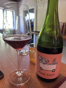 A bottle of wine that says Road Soda and a wine glass with red wine in it.
