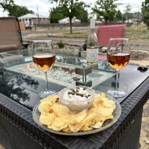 a dip surrounded by chips and topped with capers. Two glasses of rose wine are behind the plate.