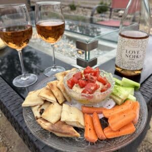 A bowl with a dip in it and tomatoes on top. On a plate with crackers and fresh veggies. With two glasses of wine on the side.