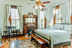 A sun-drenched room with a king bed, fireplace, several windows, wood floors, a table and chairs.