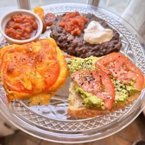 a glass dish with several food items on it including an egg item topped with tomato and cheese, avocado toast, black beans, and salsa.
