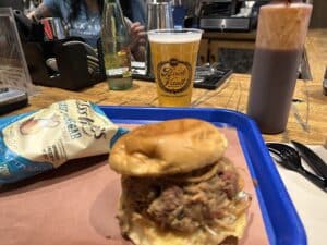 A chopped meat sandwich, a bag of chips, a squeeze bottle of BBQ sauce, and a draft beer that says Brick Vault.