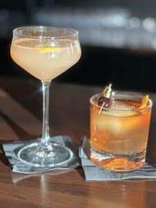 Two cocktails. One is light in color and in a champagne glass. The other is in a low glass with ice. The liquid is brownish.