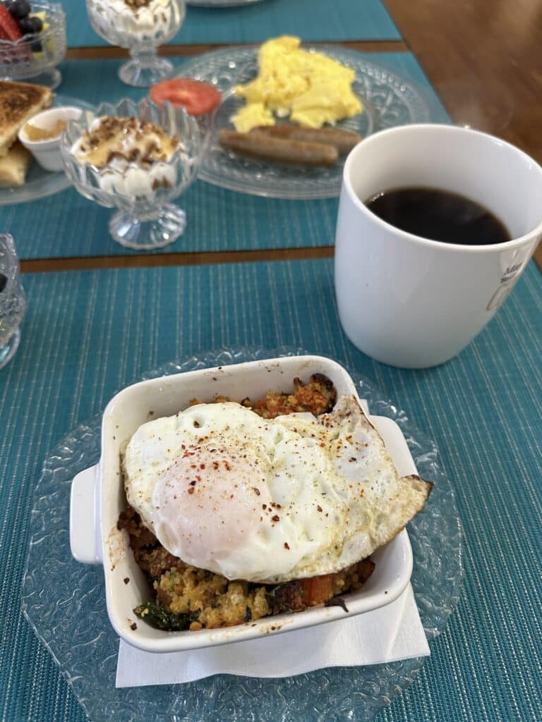 A table set with teal placemats, a white square bowl with what looks like bread and tomatoes with a fried egg on top. There's a cup of coffee and other dishes in the background
