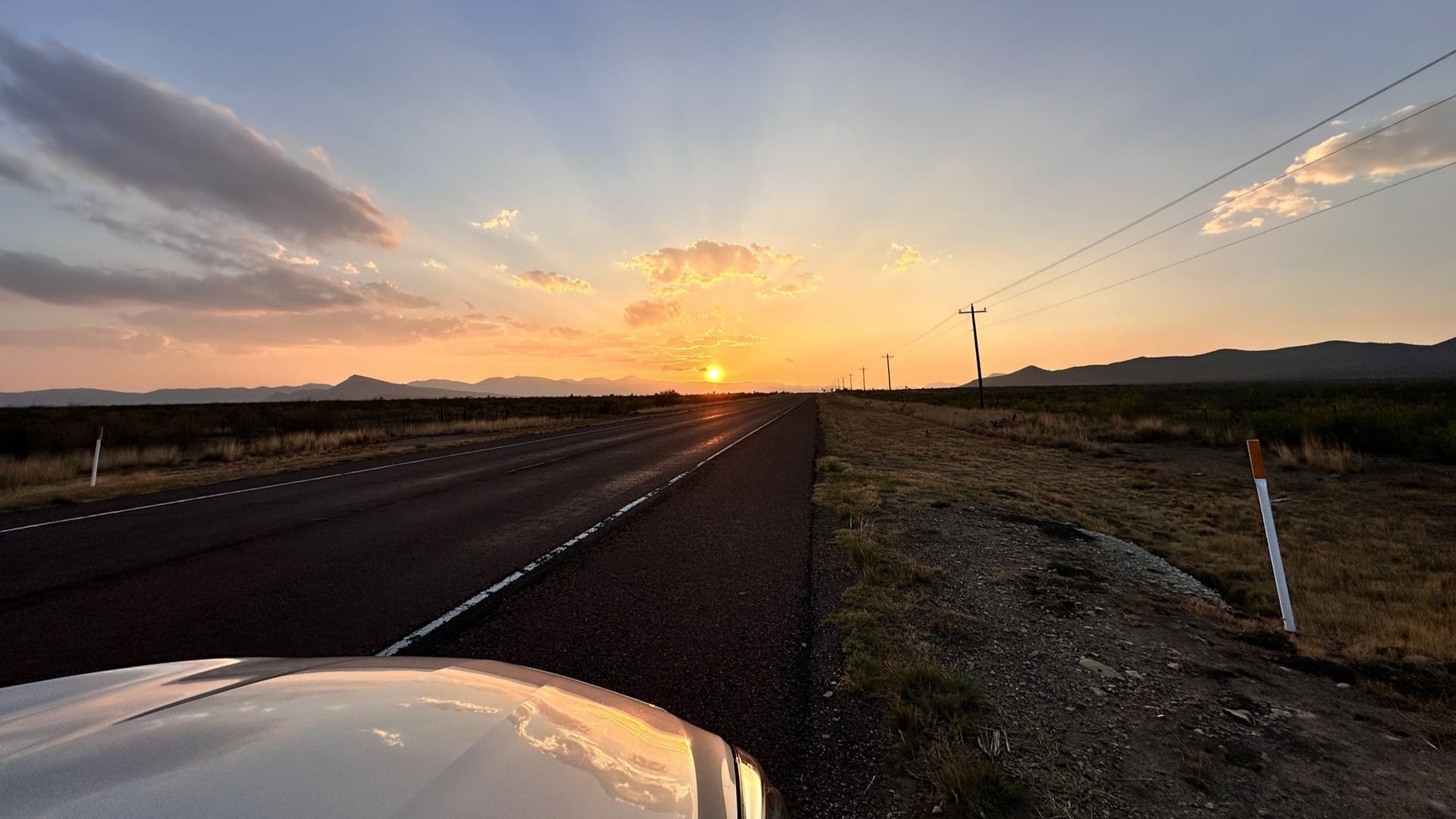 A photo of a sunset with mountains and rays coming from the sun. Part of the hood of the car is also in the picture, with a reflection of the clouds on it.