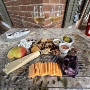 a cheese plate with 3 cheeses, apple, crackers, pickles and more, with two glasses of wine.