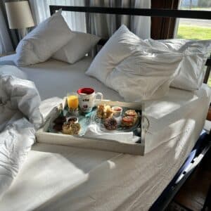 a tray of food, coffee and juice on a tray on a bed with light shining through a window.