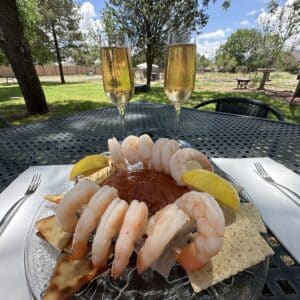 12 shrimp on a plate with lemon wedges, cocktail sauce, and crackers. Two glasses of wine are behind it.