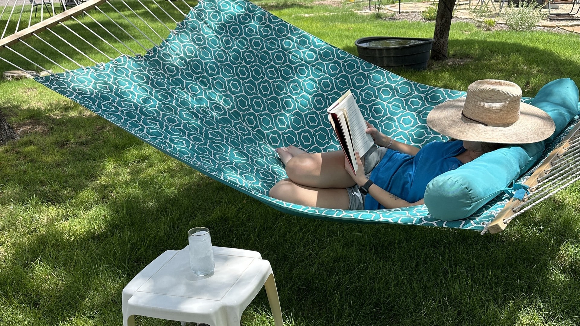 A woman lying on a hammock with a book and a floppy hat. A drink is on a table next to her. The hammock is in the shade over green grass.