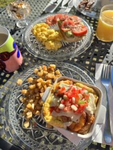 An outdoor table set with multiple plates of food, coffee, and juice. There is an egg dish, potatoes, tomatoes, yogurt, and more