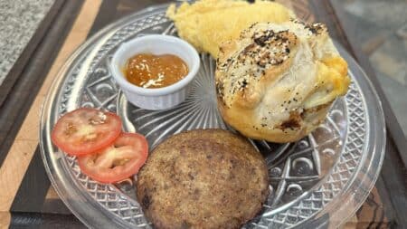 a plate of food with a small ramekin of jam, a sausage patty, a stuffed pastry with seasoning on tup, tomato slices, and a yellow item that could be scrambled eggs or some sort of grits.