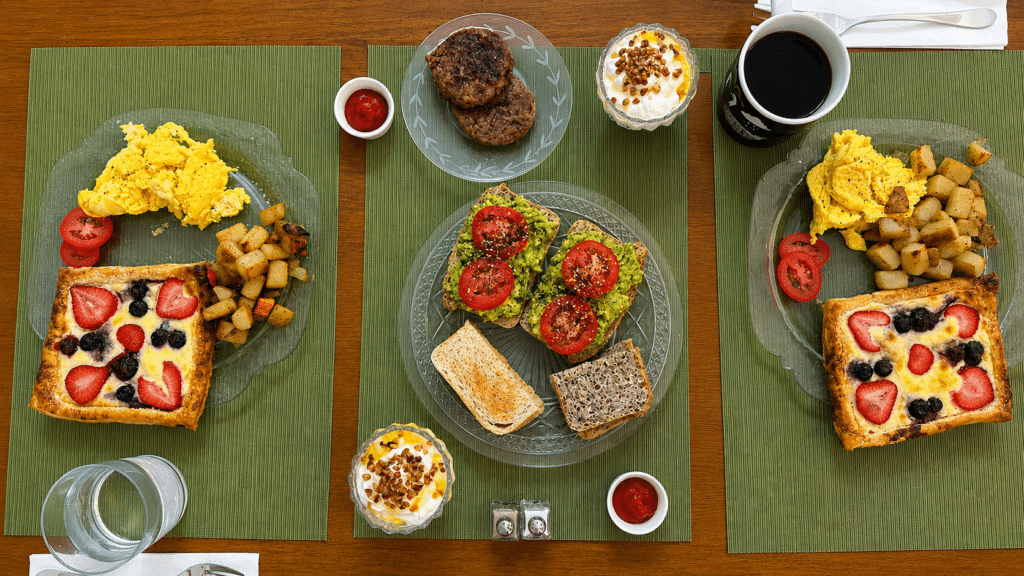 A breakfast spread on green placemats - scrambled eggs, berry danish, avocado toast, regular toat, yogurt with honey and pecans, potatoes, sausage, and coffee.