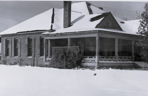 A black and white historic photograph of a brick home with a front porch and snow on the ground
