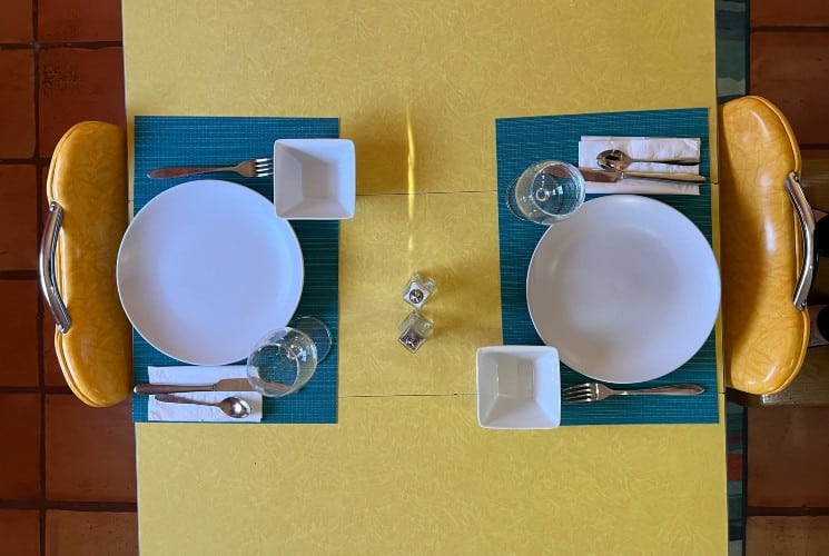 A yellow breakfast table with two chairs set for two with blue placemats and white dishes