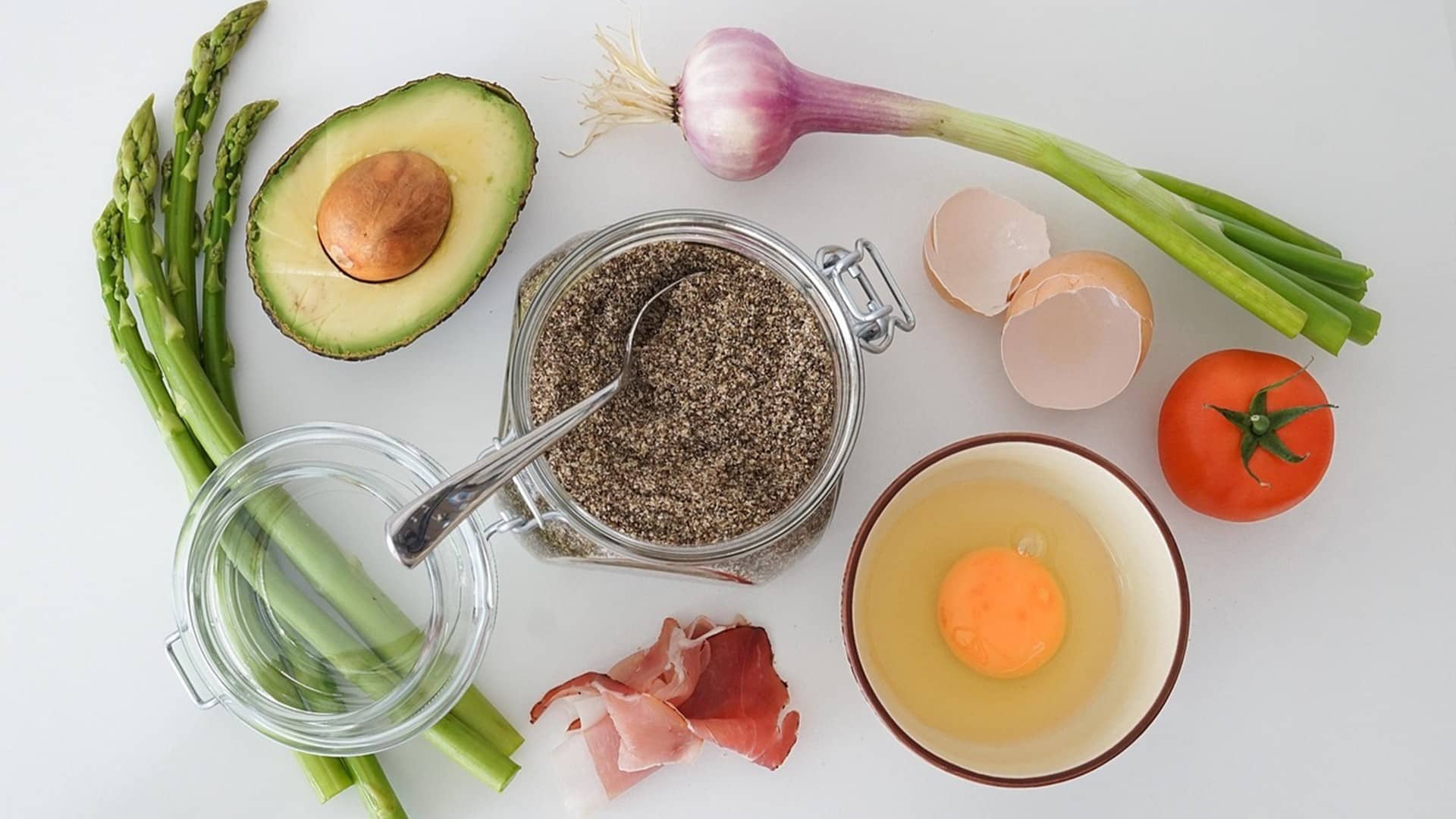 asparagus, half avocado with pit, a purple onion with its stalk, egg shels, a tomato, an egg in a bowl, a slice of Prosciutto, and a glass canister of black pepper artfully arranged on a white counter.