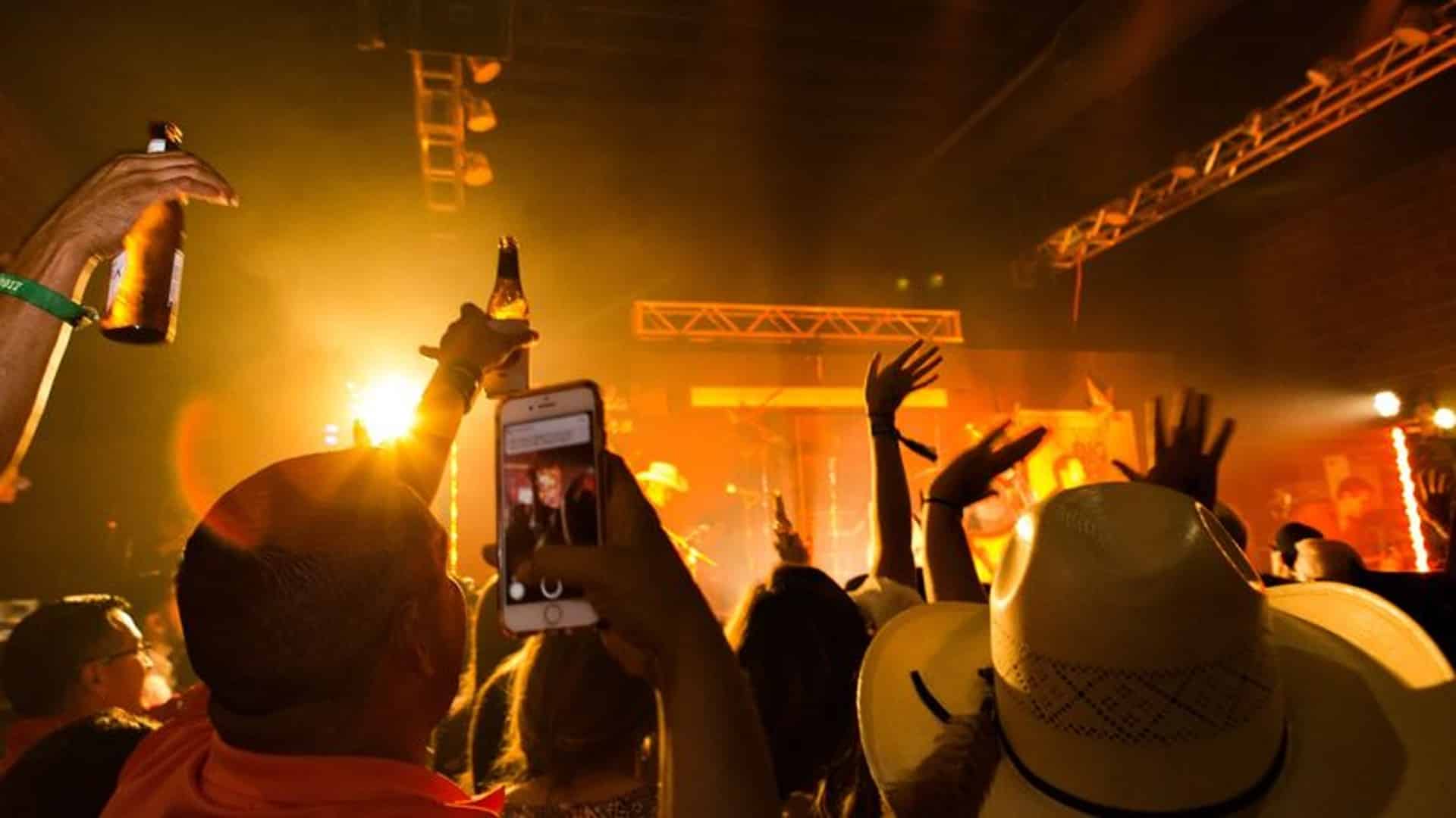 View of a crowd at night with hands raised by a stage with a band playing under golden lights