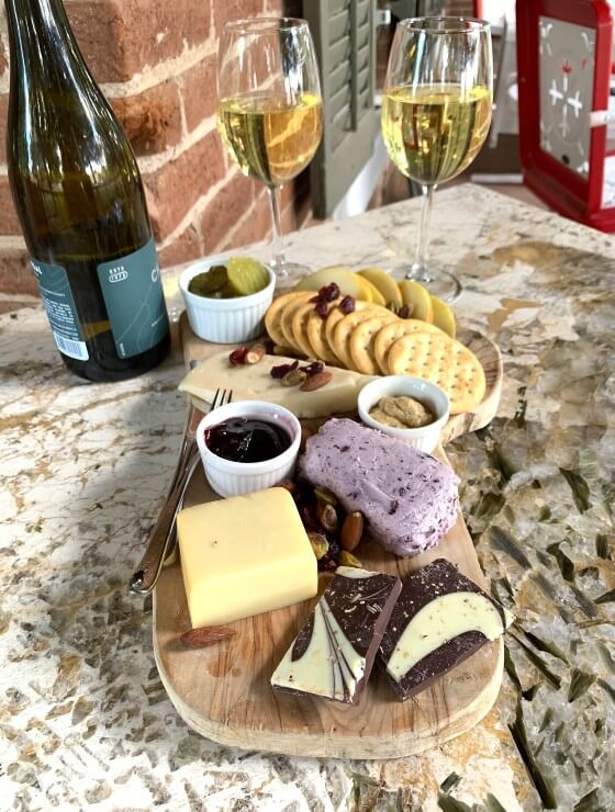 Wood platter charcuterie board of crackers, chocolate, nuts and cheeses with two glasses and a bottle of wine