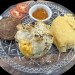 a plate of food with a small ramekin of jam, a sausage patty, a stuffed pastry with seasoning on tup, tomato slices, and a yellow item that could be scrambled eggs or some sort of grits.