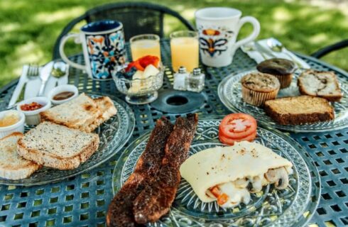 Outdoor round table with several glass plates with breakfast foods, cups of orange juice, and two mugs of coffee