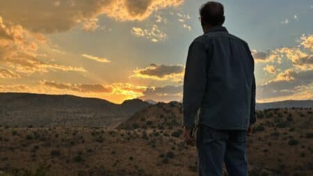 a man standing with his back to the camera, looking at the sun setting behond some mountains in the desert
