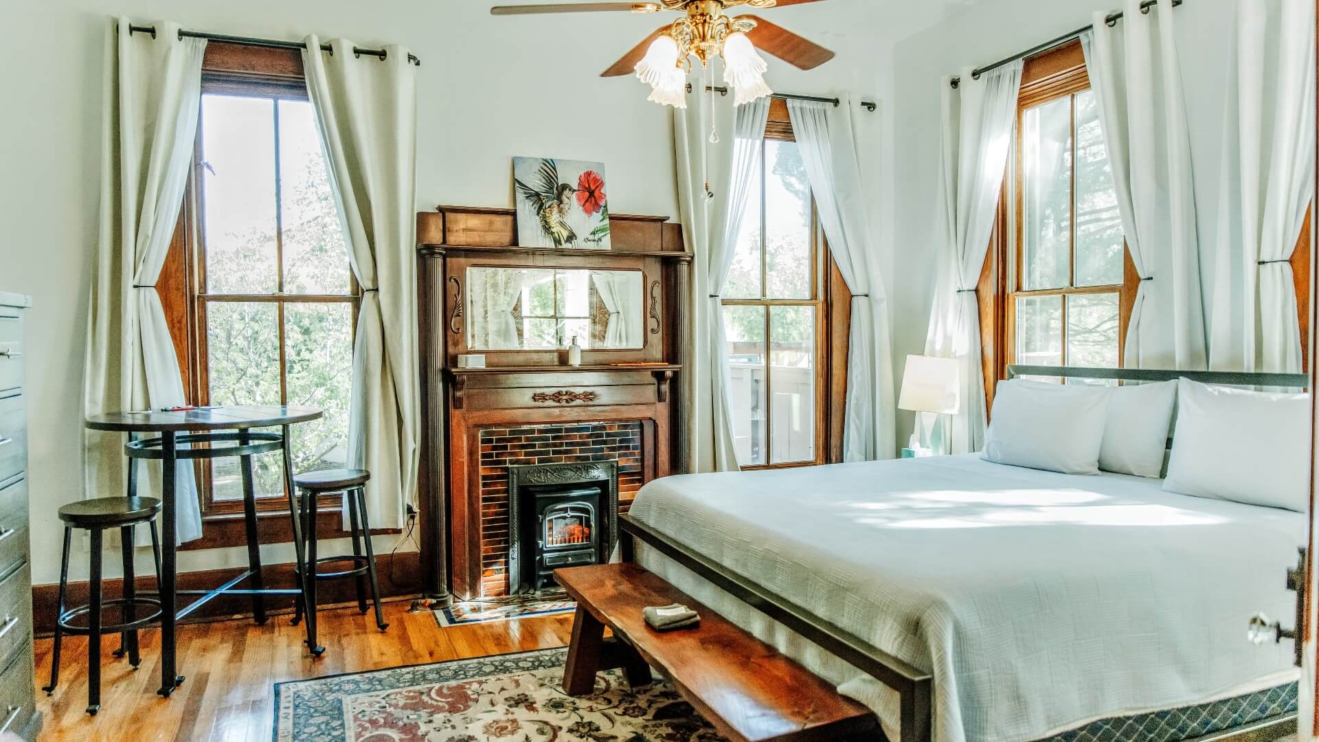 Elegant bedroom with king bed, fireplace, bistro table with two stools and large windows with white curtains