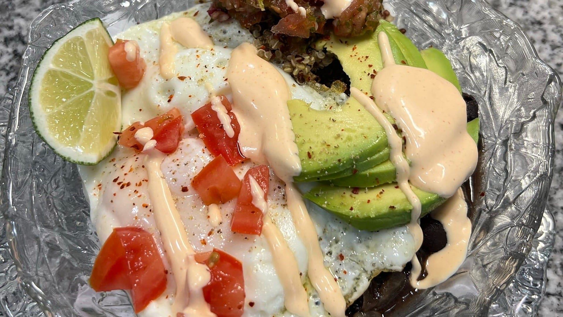 Eggs, avocado, tomato, quinoa, spicy aioli, and slice of lime on a glass plate