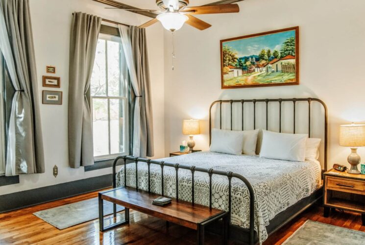 Bright bedroom with bed, side tables with lamps, large windows with grey curtains and hardwood floors