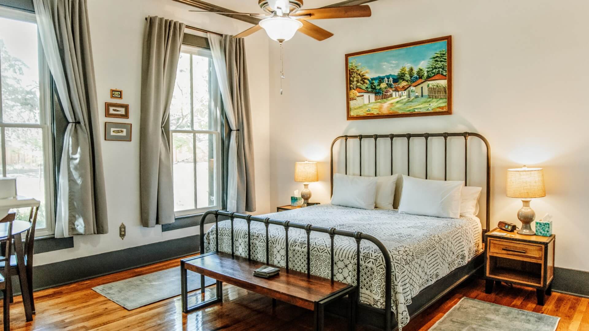 Bright bedroom with bed, side tables with lamps, large windows with grey curtains and hardwood floors