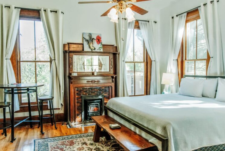 Elegant bedroom with king bed, fireplace, bistro table with two stools and large windows with white curtains
