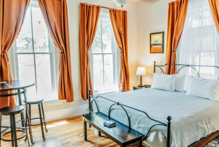 Bedroom with king bed, bistro table with two stools and large bright windows with rust colored curtains