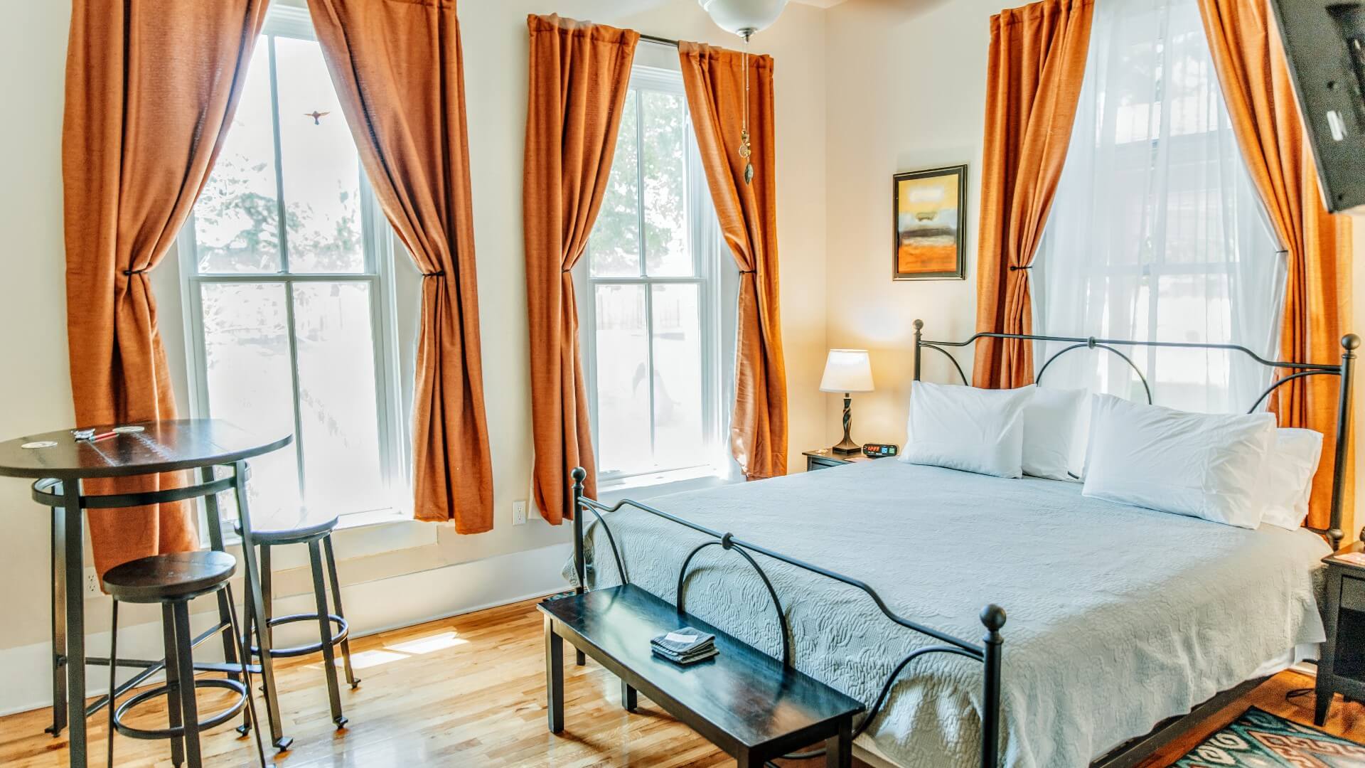 Bedroom with king bed, bistro table with two stools and large bright windows with rust colored curtains